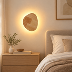 The asymmetrical panel displays visible natural-looking grain, lending a subtle handcrafted mood to a modern room. Rated at 12W, 1100 lumens and 2700K, the fixture creates a warm ambient accent around beds, sofas and quiet corners. 